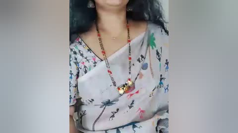 Media: Video of a woman with long black hair, wearing a colorful floral saree with a gold necklace featuring a small pendant, against a blurred background.