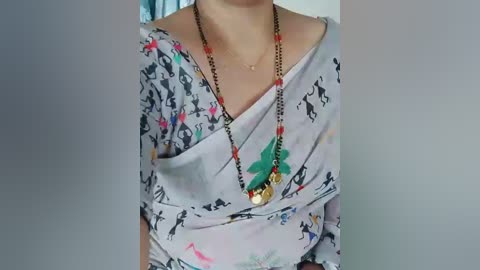 Video of a person wearing a light-colored, patterned dress with a black shoulder strap, adorned with a long, beaded necklace featuring green, red, and gold beads. Background is blurred.