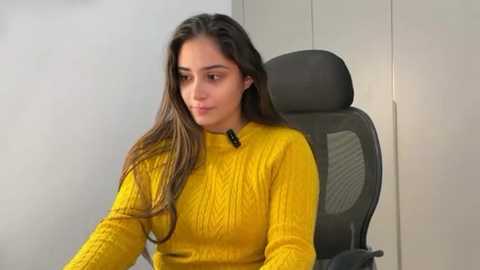 Video of a young woman with long, straight brown hair, wearing a mustard-yellow, ribbed-knit sweater, seated in a black office chair against a plain white wall.