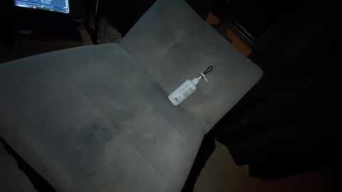 Video of a dimly lit room with a white, rectangular object on a gray, textured mat. The object, resembling a keychain, is centrally placed on the mat. Background includes dark furniture and a partially visible computer screen displaying a digital interface.