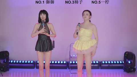 Video of two Asian women singing on stage; one in a black leather dress, the other in a yellow strapless dress. Background: white wall with Chinese text, blue LED lights, and stage lighting.