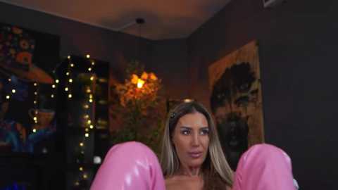 Video of a woman with long blonde hair, wearing pink gloves, standing in a dimly-lit room with dark walls, string lights, and colorful artwork.