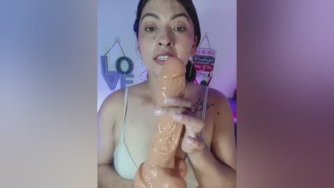 A video of a Latina woman with medium skin tone, dark hair, and a light blue bra, holding a large, realistic flesh-colored dildo, against a backdrop of wall decor with inspirational signs.
