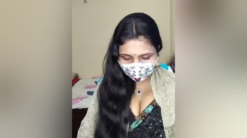 Video of a woman with long black hair, wearing a face mask and a grey cardigan over a black dress, sitting in a small room with a bed and pink and white bedding in the background.