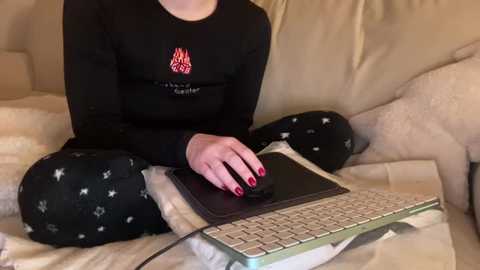 Video of a person with red nails, wearing black long-sleeve shirt and pajama pants with stars, typing on a laptop on a beige couch.