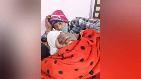 Video of an infant wrapped in a red blanket with black polka dots, lying on a gray and white plaid blanket, being held by an adult wearing a pink and blue beanie.