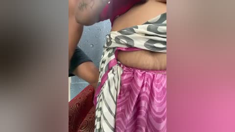 A video of a woman with light brown skin, wearing a pink and white patterned skirt and a black bra, leaning against a wall with a blue texture.