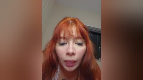Video of a young woman with long, vibrant red hair, pale skin, and a drooling expression. She's indoors, with a blurry background featuring a window.