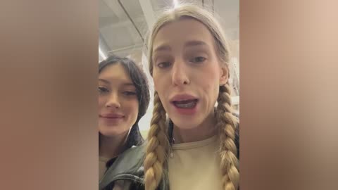 Video of two young women with light skin, one with braided blonde hair and the other with dark hair, smiling in a casual indoor setting.