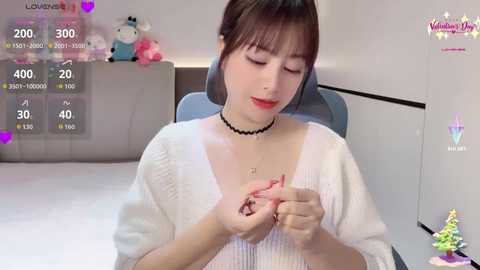 Video of a young Asian woman with straight brown hair, fair skin, wearing a white knitted cardigan, black choker, and red lipstick, sitting in a modern living room, holding a small object.