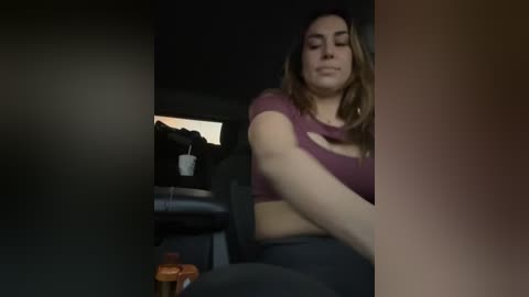 Video of a woman with long, light brown hair, wearing a burgundy crop top and black leggings, sitting in a dark car with a water bottle beside her.