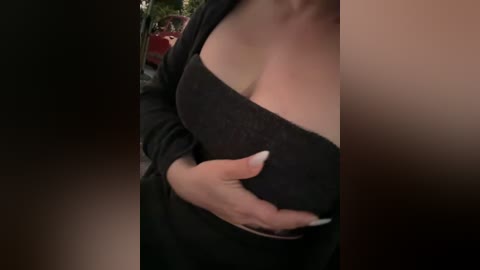 A close-up video of a woman with light skin, wearing a black off-shoulder top and a black cardigan, revealing her cleavage. She has a hand on her chest. Background shows blurred outdoor scene with a red car.
