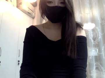 Video of an Asian woman with long, straight black hair, wearing a black mask, black off-the-shoulder top, and a white bag. She stands in a brightly lit room with white walls and a patterned curtain in the background.