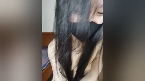 Media: A video of a woman with long black hair, partially obscured by a black mask, wearing a beige sweater, seated on a wooden bench. The background shows a blurred, neutral-colored wall.