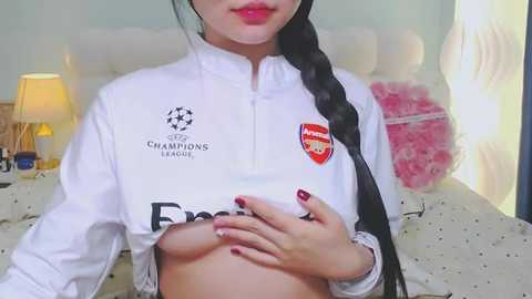 A video of an Asian woman with long black braided hair, wearing a white Arsenal football shirt, revealing her medium-sized breasts, standing in a softly lit bedroom with a pink teddy bear and a lamp.