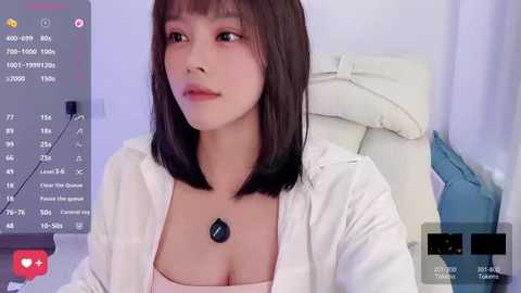 Video of a young Asian woman with straight black hair, light skin, and pink lipstick, wearing a white blouse and necklace, sitting on a white chair in a soft-lit room.