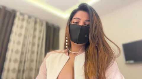 A video of a young woman with long brown hair, wearing a black face mask and a white blouse, standing indoors with beige curtains and a TV on the wall.