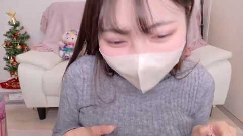 Video of a young Asian woman with straight, shoulder-length dark hair, wearing a gray ribbed sweater and white face mask, kneeling indoors near a white sofa, decorated with a festive Christmas tree, plush toys, and a red blanket.