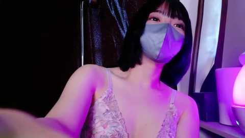Video of an Asian woman with straight black hair, wearing a light blue surgical mask and a lace-patterned white bra, standing in a dimly-lit room with purple lighting, holding a selfie stick.