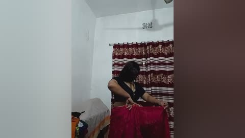 A video of a woman in a modest, dark-colored blouse and red sari, adjusting a curtain in a simple, white-walled room with maroon and white striped curtains.