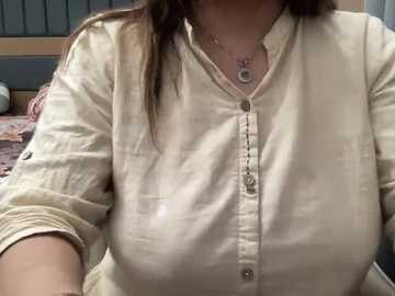 A video of a woman with medium breasts, wearing a beige button-up shirt and a necklace, seated on a bed with patterned bedding, in a dimly lit room.