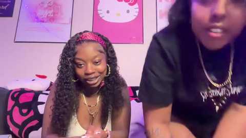 Video of two young Black women with curly hair, one wearing a red headband, sitting on a bed with pink Hello Kitty bedding, smiling.
