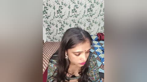 Media: Video of a South Asian woman with long dark hair, wearing a colorful saree and a red bindi, sitting on a patterned bedspread, with a white brick wall and green ivy wallpaper in the background.