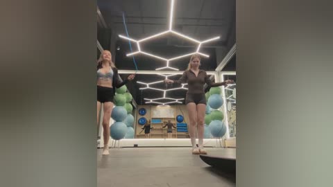 Video of two women in a modern gym with hexagonal lighting, wearing sports bras and shorts, dancing on balance balls. Background features blue and green exercise balls.