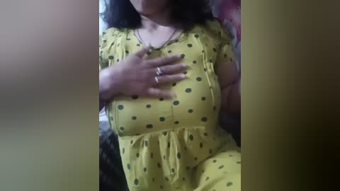Media: Video of a woman with medium skin tone, wearing a yellow dress with black polka dots, placing her hand on her chest. Background is blurred, suggesting an indoor setting.