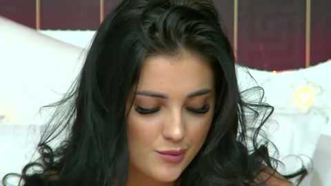 Video of a young woman with long, wavy black hair and fair skin, eyes closed, wearing subtle makeup, in a dimly lit room with white and dark maroon wallpaper.