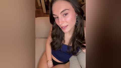 Video of a young woman with light skin and dark wavy hair, wearing a blue tank top and black shorts, sitting on a beige sofa in a warm-toned room.