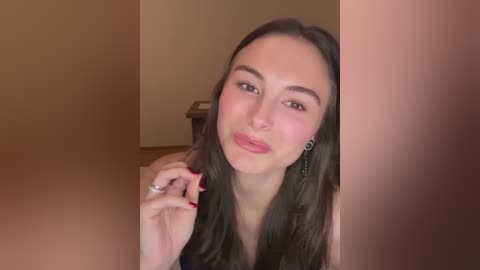 Video of a young woman with long, straight brown hair, fair skin, and pink blush on her cheeks. She wears a sleeveless top and red nail polish. Background shows a beige wall and a wooden table.