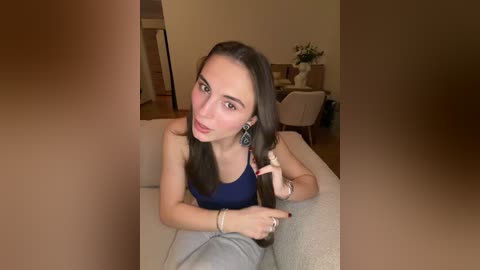A video of a young woman with fair skin, long brown hair, and a blue tank top, sitting on a beige couch in a modern living room with beige walls and a white table.
