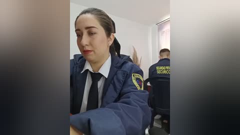 Media: Video of a serious-looking woman with dark hair in a ponytail, wearing a navy blue police uniform and tie, seated in a room with blurred background, including a person in uniform.