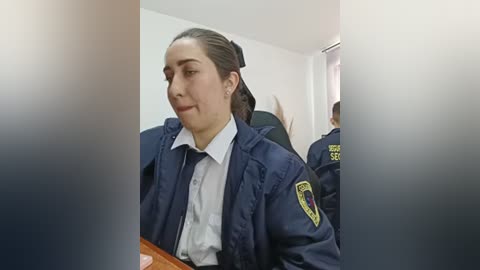 Video of a Latina woman with light skin and dark hair in a ponytail, wearing a navy blue police uniform with a yellow badge, seated in a plain white room, possibly an interrogation room.