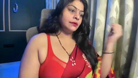 Video of a South Asian woman with long black hair, wearing a red saree with floral embroidery, seated on a beige chair, indoors.