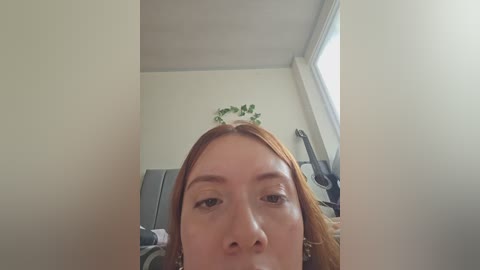 A close-up photo of a young woman with light skin and straight, reddish-brown hair, making a pouty face in a bright, modern room with white walls and a large window.