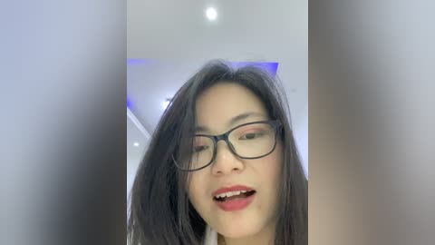 Video of an East Asian woman with straight black hair, wearing black-rimmed glasses, and a white shirt, smiling with slightly parted lips. Background shows blurred modern interior with soft lighting and white walls.