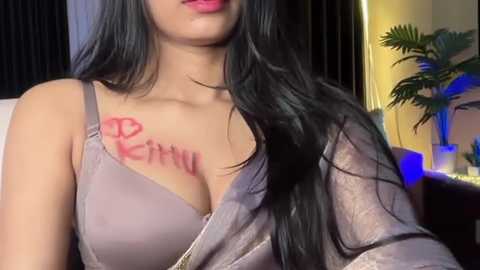 Video of a young woman with long black hair, light skin, wearing a low-cut gray tank top revealing a red \"Kitty\" tattoo on her left breast. Background shows a potted plant and dimly lit room.