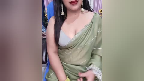 Video of a South Asian woman with long black hair and fair skin, wearing a green saree with a white bra visible, sitting indoors with a blue chair and purple curtains in the background.