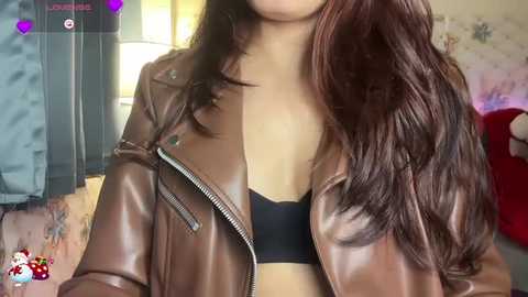 A video of a light-skinned woman with long, wavy brown hair, wearing a black bra and a brown leather jacket. The background features a blurred, indoor setting with floral wallpaper.