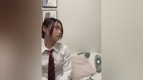 Video of an Asian woman with dark hair tied back, wearing a white shirt and red tie, sitting on a white couch with green patterned cushions. She looks thoughtful against a plain white wall with framed art.