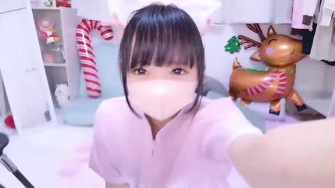 Video of an Asian woman with short black hair, wearing a pink surgical mask, taking a selfie in a festive room with Christmas decorations, including a reindeer statue and candy canes.
