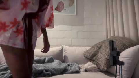 A video showing a person in a floral robe, standing in a cozy living room with a beige couch, white brick wall, and a framed art piece.