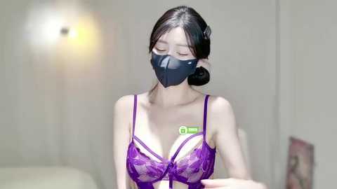 Video of an East Asian woman in a purple lace bra, wearing a black face mask, with a green QR code on her chest. She has dark hair and is indoors with blurred background.