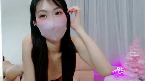 Video of an East Asian woman with long black hair, wearing a pink mask, blowing a bubble, in a pink room with white curtains.