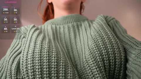 A close-up video of a fair-skinned woman with red hair wearing a chunky, textured, mint-green knitted sweater. Background is a muted beige wall.