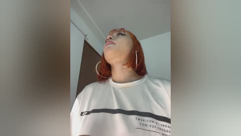 A video of a young woman with shoulder-length, straight red hair, wearing a white T-shirt with black text and large hoop earrings. She gazes upward with a neutral expression. The background is a plain, white-walled room.