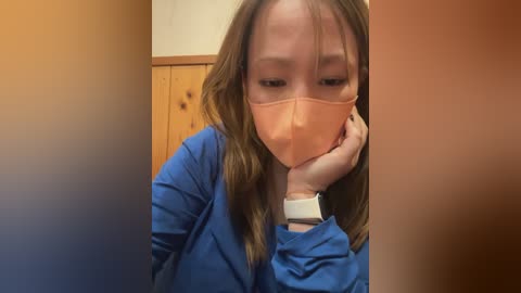 Video of a young woman with light skin, wearing a blue shirt and a peach face mask, covering her mouth and nose. She has shoulder-length brown hair and is in a wooden room, looking down with her hand on her cheek.
