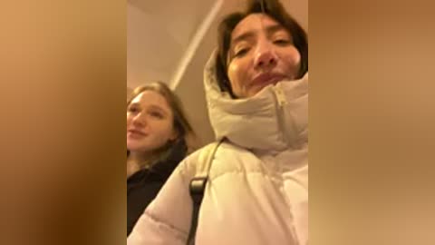 Video of two women, one in a puffy jacket, the other in a black coat, walking in a dimly lit hallway.